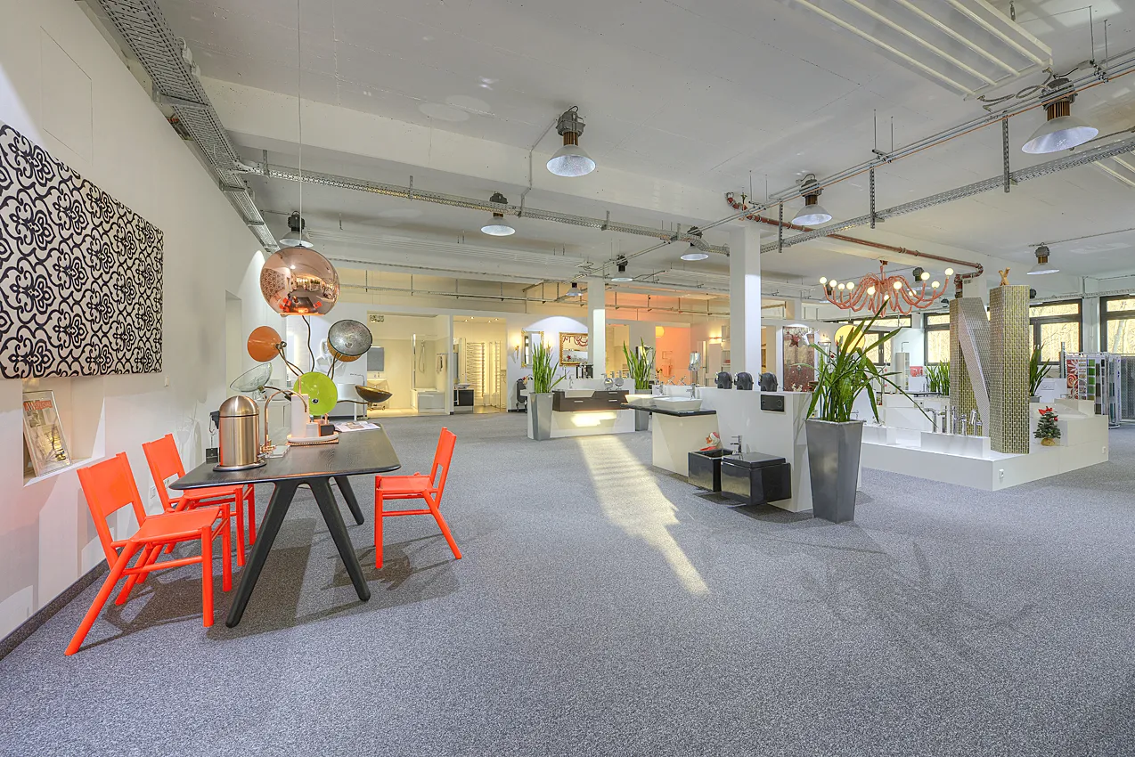 Ein weitläufiger Ausstellungsbereich der Nessmann bad & heizung GmbH mit modernen Waschtischen, Design-Elementen, auffälligen Leuchten und einem stilvollen Besprechungsbereich mit orangenen Stühlen.