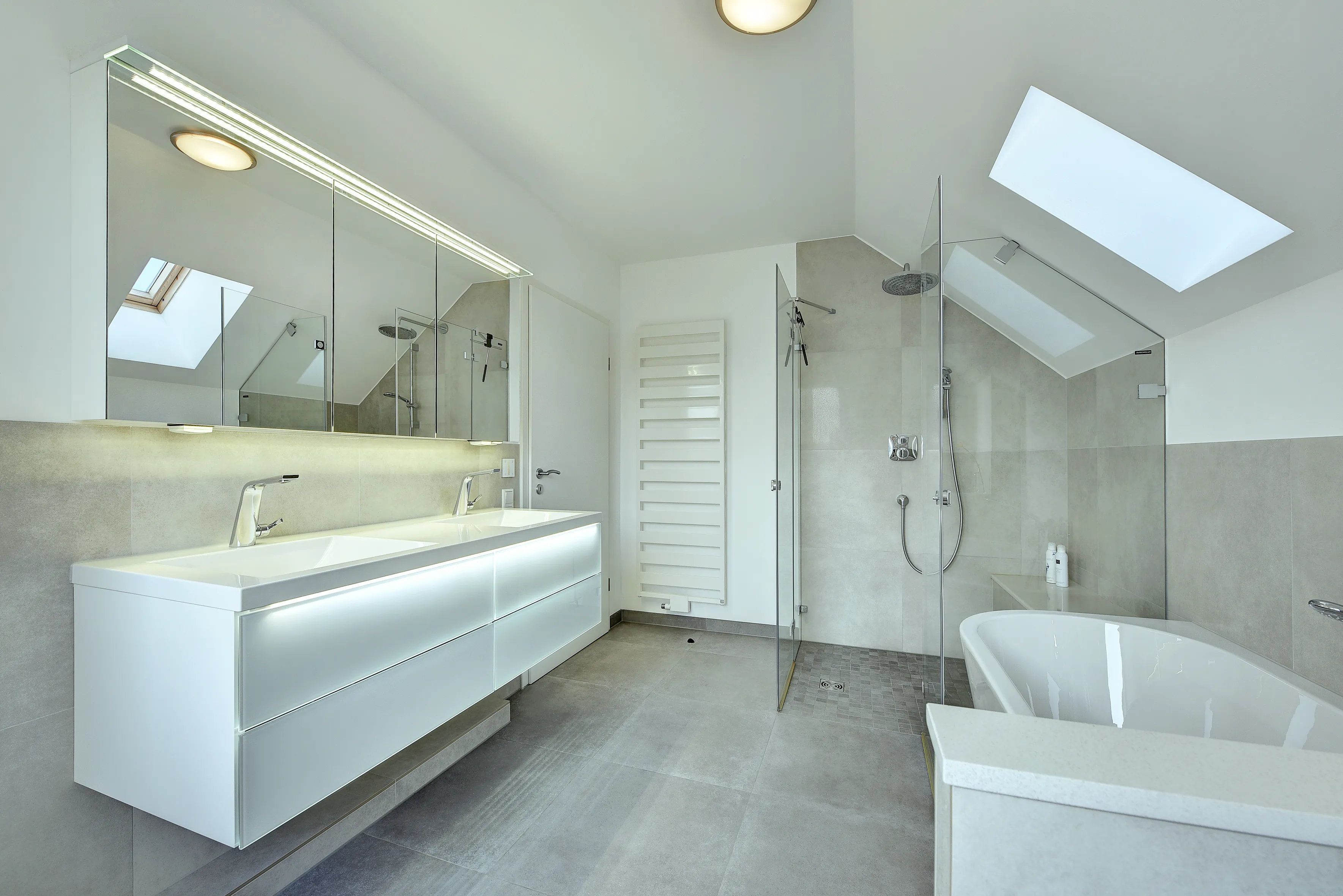 Modernes Dachbad mit beleuchtetem Waschtisch, Glasdusche und Badewanne in hellen Naturtönen, umgesetzt von der Nessmann Bad & Heizung GmbH.