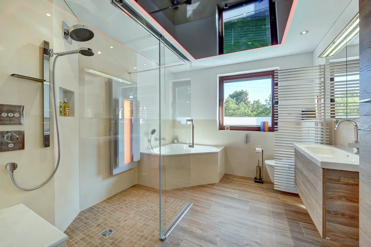 Modernes, helles Badezimmer mit bodengleicher Glasdusche, Badewanne, Holzwaschtisch und LED-Deckenbeleuchtung, realisiert von der Nessmann Bad & Heizung GmbH.