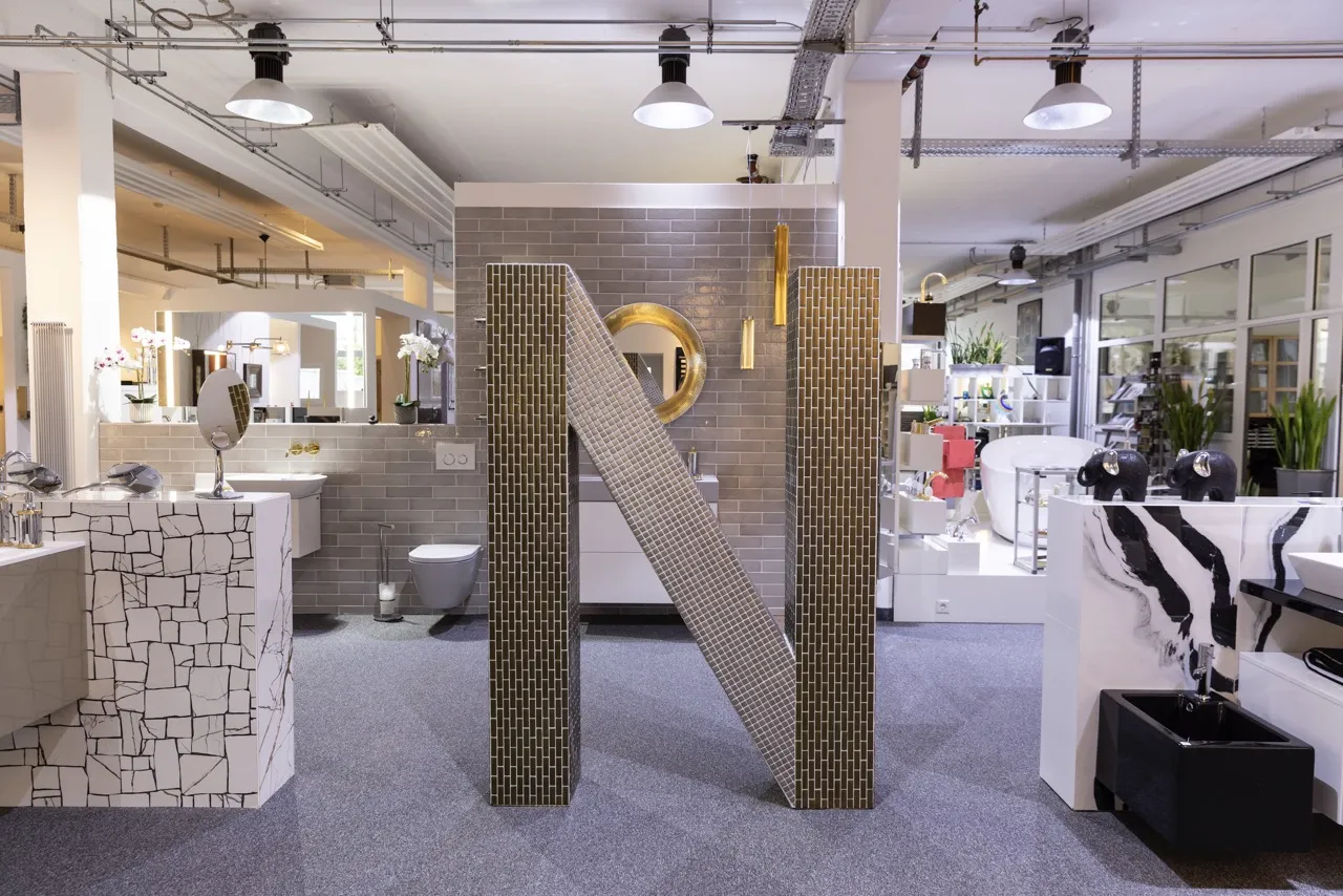 Ein stilvoll gestalteter Ausstellungsbereich der Nessmann bad & heizung GmbH mit modernen Waschtischen, goldenen Akzenten und einem großen, mit Fliesen verkleideten "N" als Designelement.
