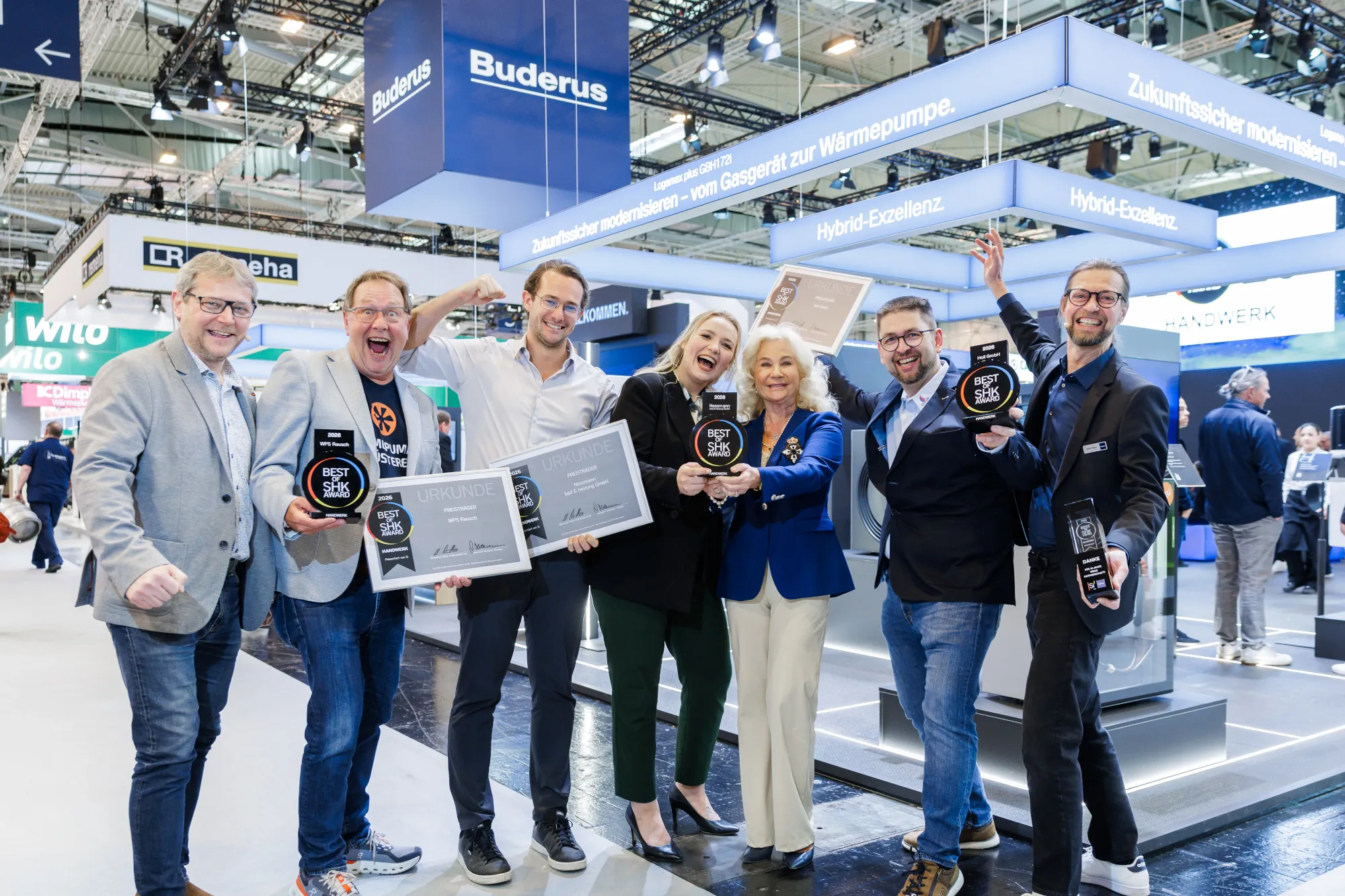 Eine Gruppe von sieben Personen jubelt auf einer SHK-Fachmesse vor einem großen Buderus-Messestand und präsentiert mehrere „Best of SHK Award 2026“-Trophäen sowie gerahmte Urkunden.