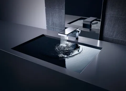 Eine hochwertige Waschtisch-Armatur, aus der Wasser in ein rechteckiges, glänzendes Waschbecken fließt..