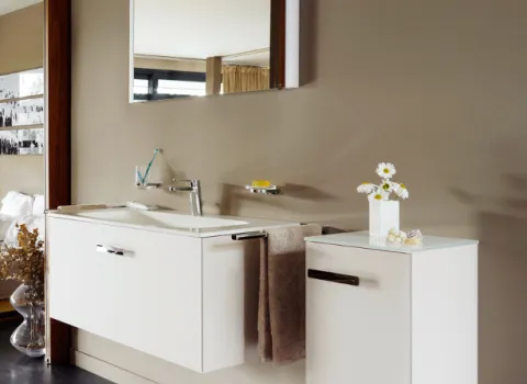 Ein modernes Badezimmer mit einem weißen Waschbecken, das an einer glatten, beige gestrichenen Wand montiert ist. Ein großer Spiegel hängt über dem Waschbecken, das von minimalistischen Schränken flankiert wird, auf denen eine Vase mit weißen Gänseblümchen steht. Die Oberflächen sind makellos und spiegeln die professionelle Arbeit von Malern und Trockenbauern wider, die eine elegante und ruhige Atmosphäre geschaffen haben.