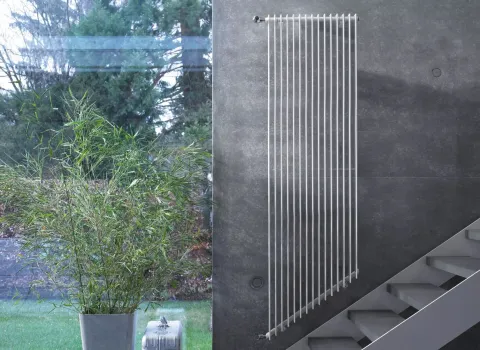 Moderner vertikaler Designheizkörper von Zehnder an dunkler Wand neben Treppe, bodentiefem Fenster und Pflanze im minimalistischen Wohnraum.