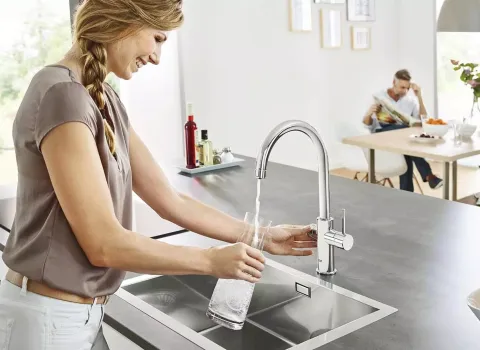 Frau zapft gefiltertes Trinkwasser aus der Grohe Blue Home Küchenarmatur in moderner Küche.