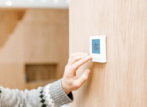 Ein Mann bedient ein digitales Thermostat, um die Heizungssteuerung im Raum anzupassen. Die Anzeige des Geräts zeigt eine Temperatur von 20°C. Die Szene spielt in einem hellen, modernen Zuhause mit Holzelementen.