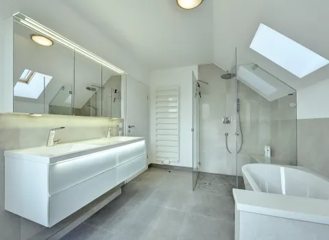 Modernes Dachbad mit beleuchtetem Waschtisch, Glasdusche und Badewanne in hellen Naturtönen, umgesetzt von der Nessmann Bad & Heizung GmbH.