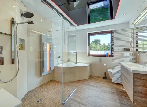 Modernes, helles Badezimmer mit bodengleicher Glasdusche, Badewanne, Holzwaschtisch und LED-Deckenbeleuchtung, realisiert von der Nessmann Bad & Heizung GmbH.