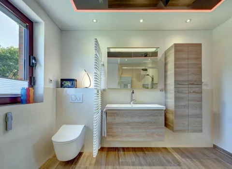 Modernes Badezimmer mit wandhängendem WC, Holzwaschtisch, Spiegelschrank, Handtuchheizkörper und indirekter LED-Deckenbeleuchtung der Nessmann Bad & Heizung GmbH.