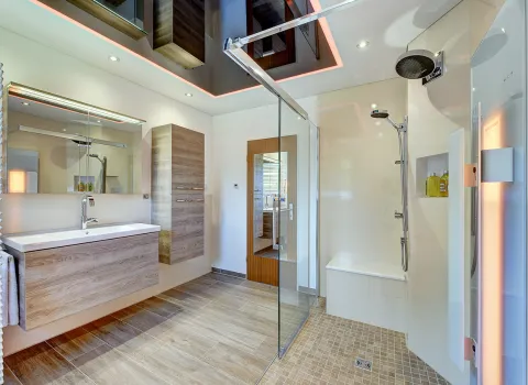 Helles Badezimmer mit bodengleicher Glasdusche, Holzwaschtisch, Hochschrank und abgehängter Decke mit LED-Lichtakzent von der Nessmann Bad & Heizung GmbH.