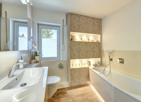 Helles Badezimmer mit Waschtisch, WC, Badewanne und beleuchteten Wandnischen, umgesetzt von der Nessmann Bad & Heizung GmbH.
