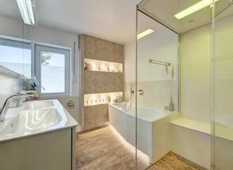 Großzügiges Badezimmer mit bodengleicher Glasdusche, Badewanne, Waschtisch und warmen Lichtakzenten, umgesetzt von der Nessmann Bad & Heizung GmbH.