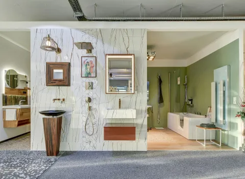 Eine stilvolle Badausstellung der Nessmann bad & heizung GmbH mit modernen Waschtischen, einer Dusche mit Glastrennwand und einer Badewanne in einem harmonischen Designmix aus Holz, Grün und Marmoroptik.