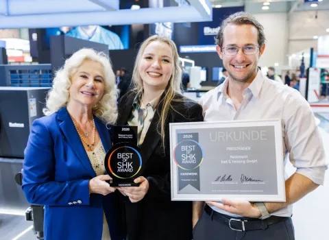 Drei Personen stehen lächelnd auf einer SHK-Fachmesse und präsentieren eine schwarze „Best of SHK Award 2026“-Trophäe sowie eine gerahmte Urkunde.