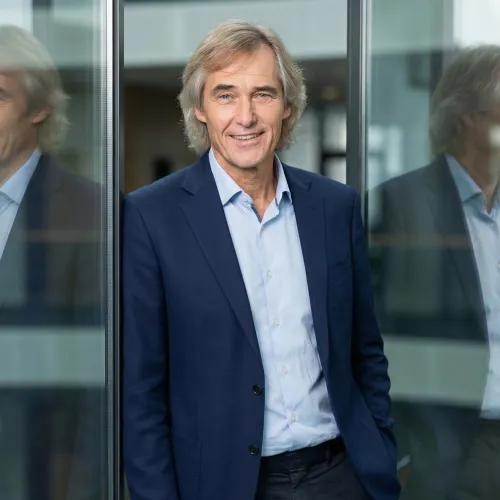 Axel Nessmann – Ein selbstbewusster Geschäftsmann mittleren Alters mit grauem Haar und blauem Sakko lächelt professionell in einem modernen Büroumfeld. Er lehnt an einer Glaswand, sein Spiegelbild ist sichtbar, was seine Präsenz unterstreicht.