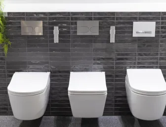 Drei moderne, wandhängende WCs mit unterschiedlichen Spülkasten-Drückerplatten sind vor einer glänzenden, dunkelgrauen Fliesenwand in der Badausstellung in Düsseldorf der Nessmann bad & heizung GmbH zu sehen.