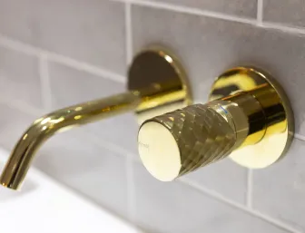 Eine luxuriöse, goldene Wandarmatur von Nessmann bad & heizung GmbH mit strukturiertem Griff und gebogenem Auslauf vor einer modernen, grauen Fliesenwand.