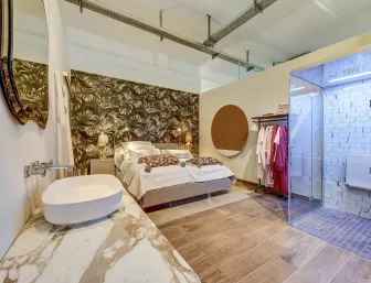 Ein stilvolles Schlafzimmer-Badezimmer-Ensemble mit einer tropisch gemusterten Tapete, einem gemütlichen Bett und einer modernen, offenen Dusche mit Glasabtrennung, präsentiert von Nessmann bad & heizung GmbH in der Badausstellung Düsseldorf.