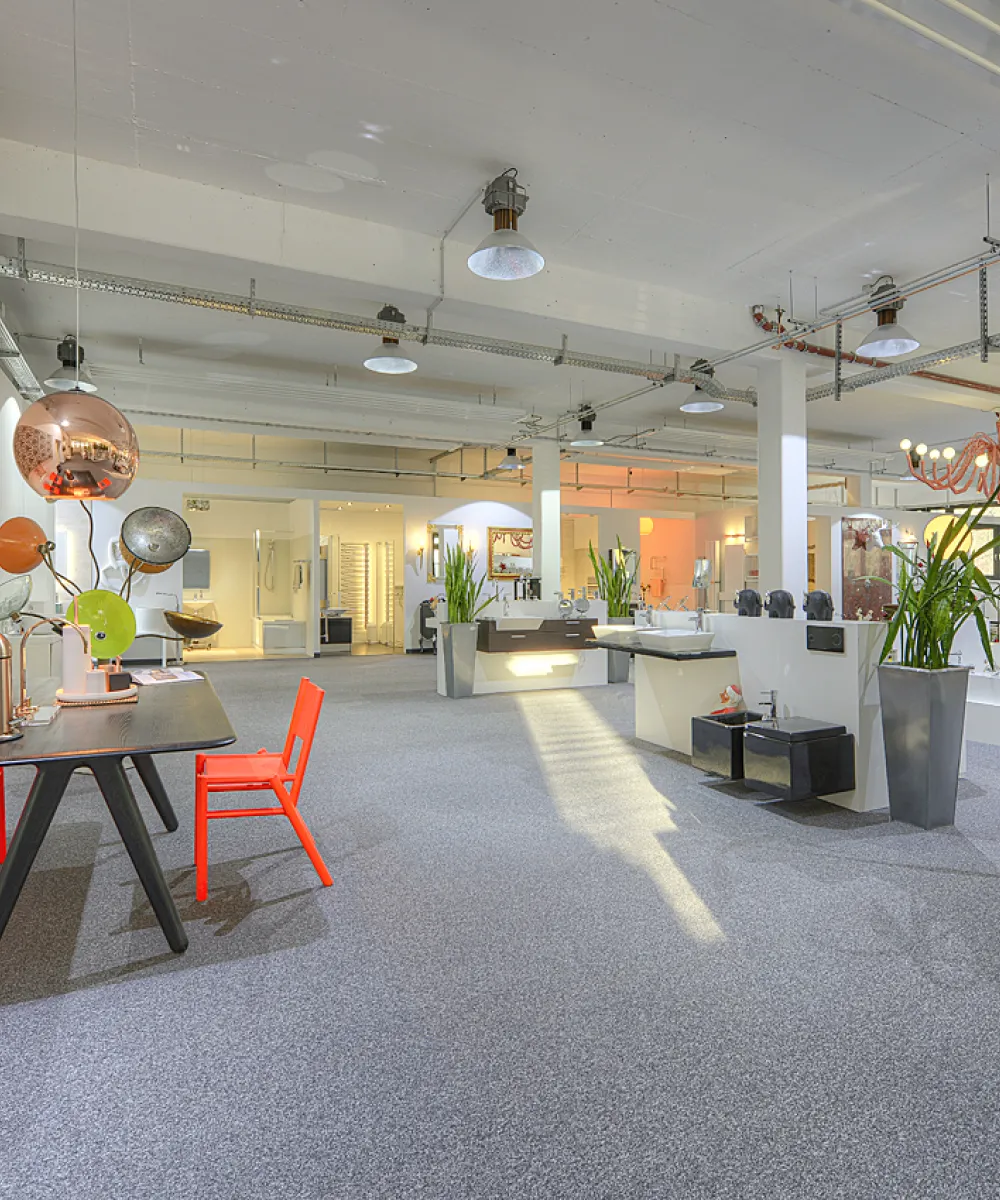 Ein weitläufiger Ausstellungsbereich der Nessmann bad & heizung GmbH mit modernen Waschtischen, Design-Elementen, auffälligen Leuchten und einem stilvollen Besprechungsbereich mit orangenen Stühlen.