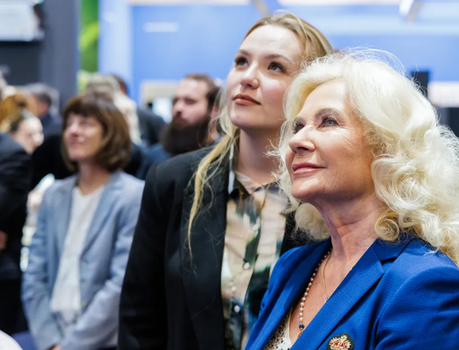 Zwei Frauen stehen im Vordergrund einer SHK-Fachveranstaltung und blicken aufmerksam nach oben, als würden sie einer Präsentation oder Preisverleihung folgen.