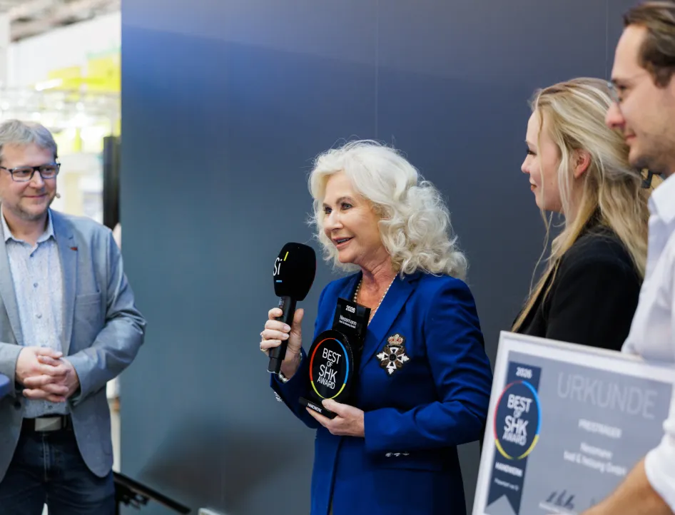 Eine Frau im blauen Blazer hält bei einer SHK Preisverleihung ein Mikrofon und eine schwarze „Best of SHK Award 2026“-Trophäe in der Hand.