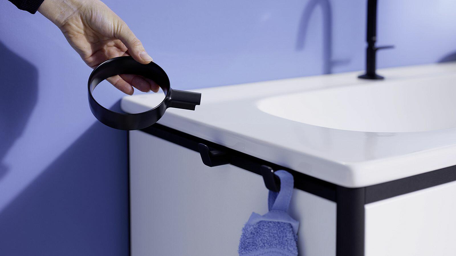 Hand mit burgbad Vry-Accessoire zeigt auf weißen Waschtisch mit schwarzen  Alurahmen. gbad